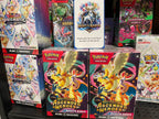 A retail display of Pokémon booster bundles, with two 'Ascended Heroes' booster bundles in the foreground and other card game boxes including Prismatic Evolutions.
