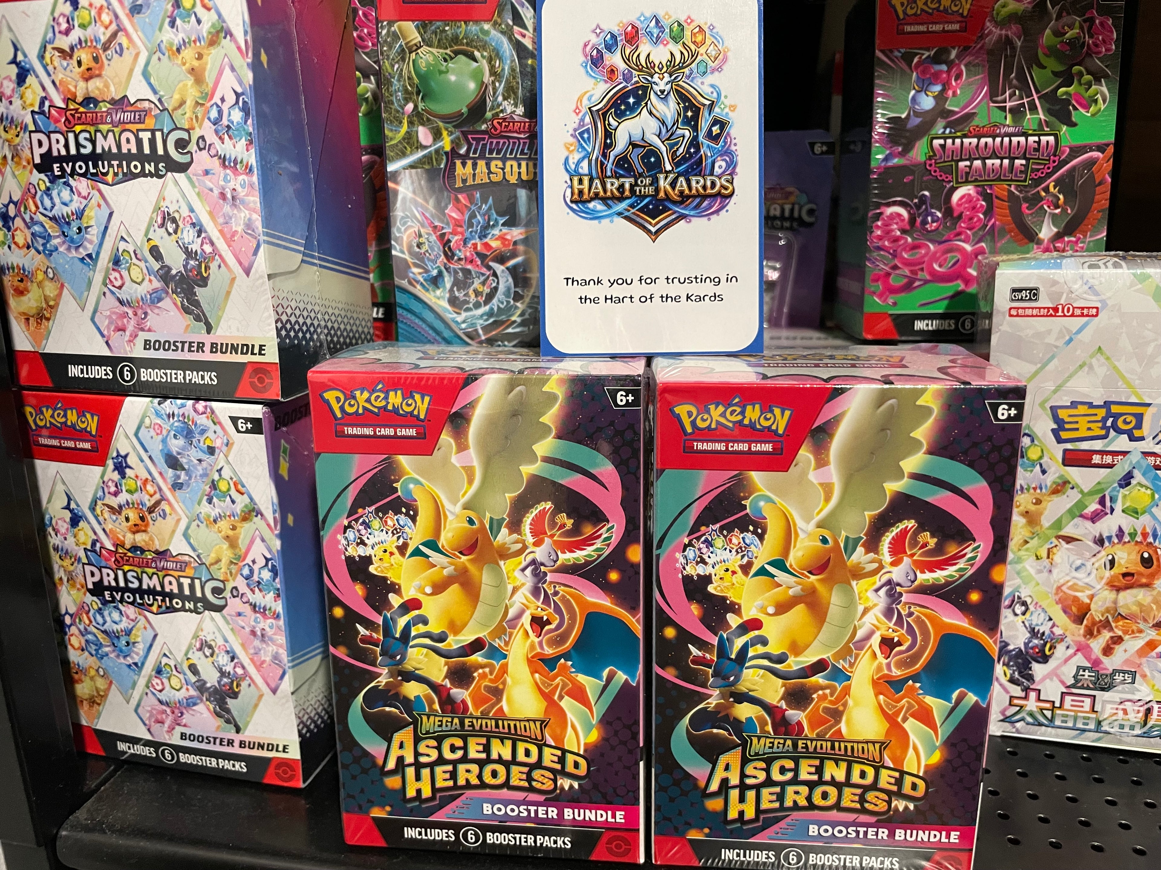 A retail display of Pokémon booster bundles, with two 'Ascended Heroes' booster bundles in the foreground and other card game boxes including Prismatic Evolutions.