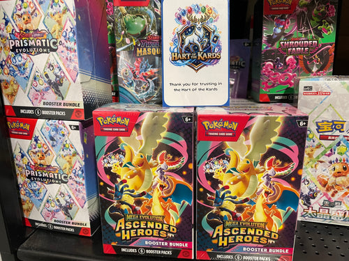 A retail display of Pokémon booster bundles, with two 'Ascended Heroes' booster bundles in the foreground and other card game boxes including Prismatic Evolutions.