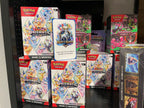 A shelf display of Pokémon TCG products, including multiple Prismatic Evolutions booster packs, all branded with the Pokémon logo.
