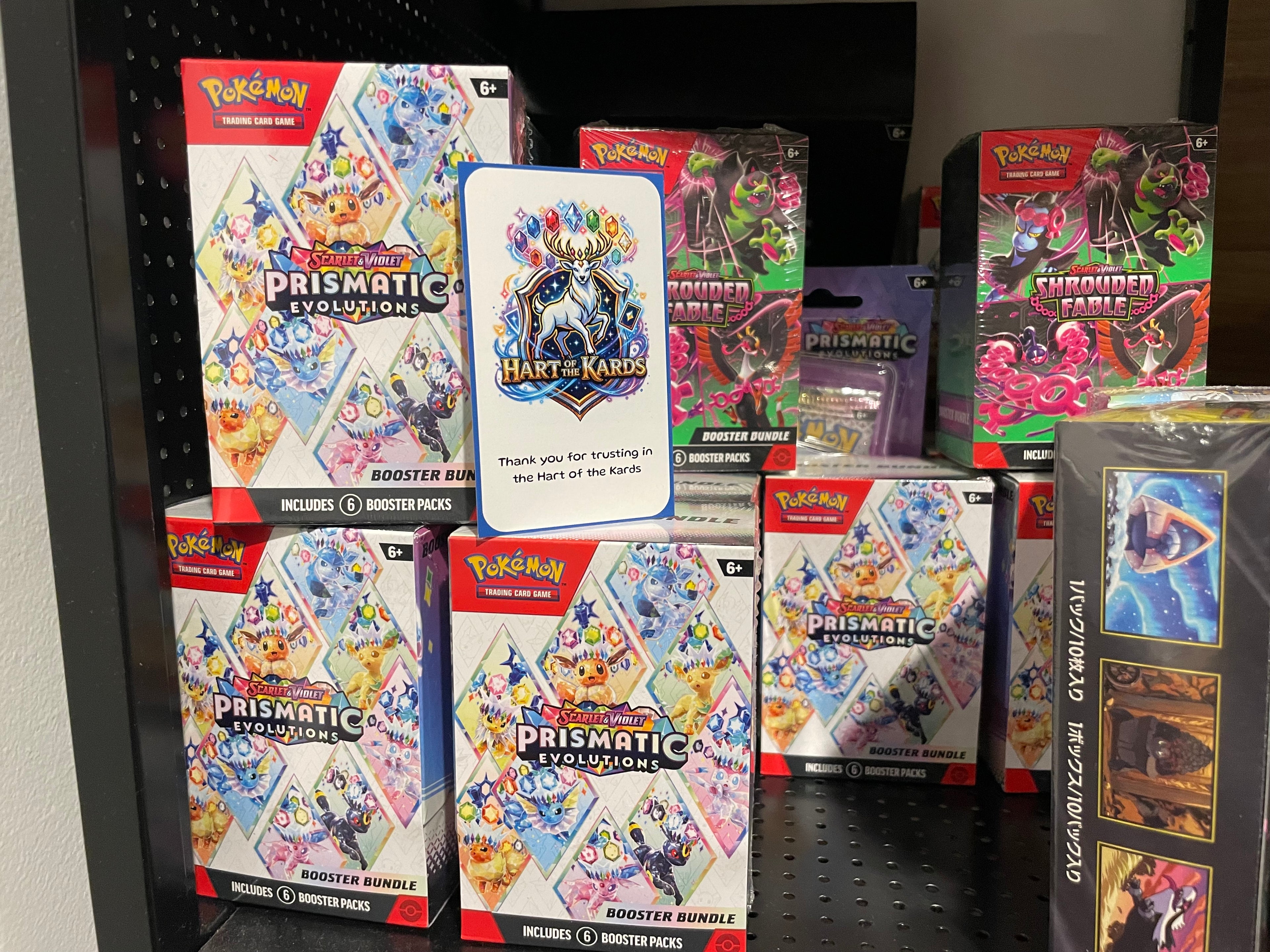 A shelf display of Pokémon TCG products, including multiple Prismatic Evolutions booster packs, all branded with the Pokémon logo.